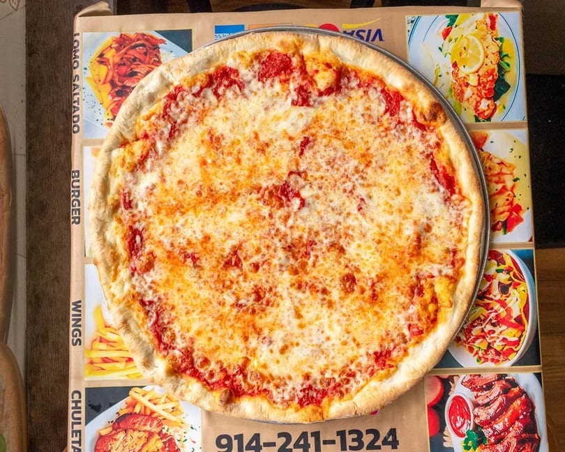 A large, cheesy pizza on a metal pan, sitting on a brown box with a menu and phone number.