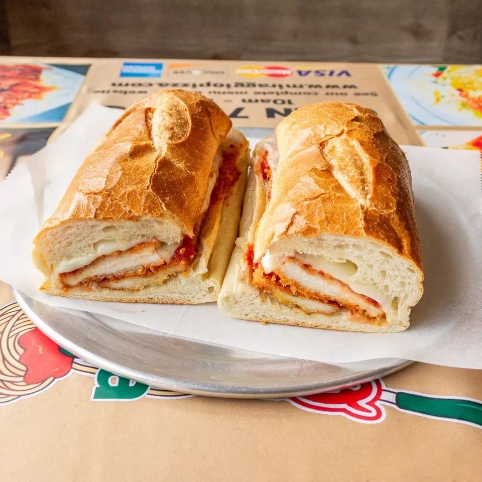 Chicken parmesan hero sandwich, cut in half, on a metal plate over a branded paper placemat.