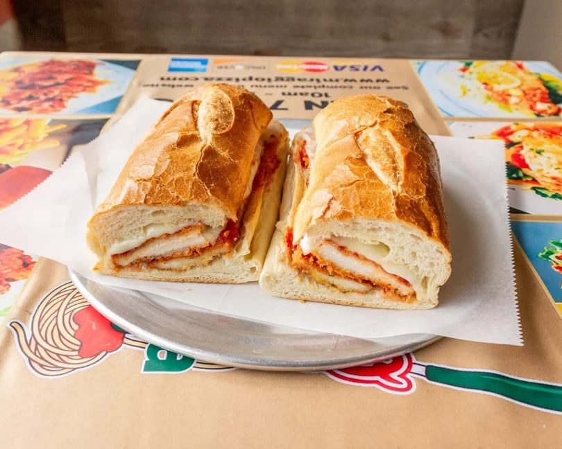 Chicken parmesan hero sandwich, cut in half, on a metal plate over a branded paper placemat.