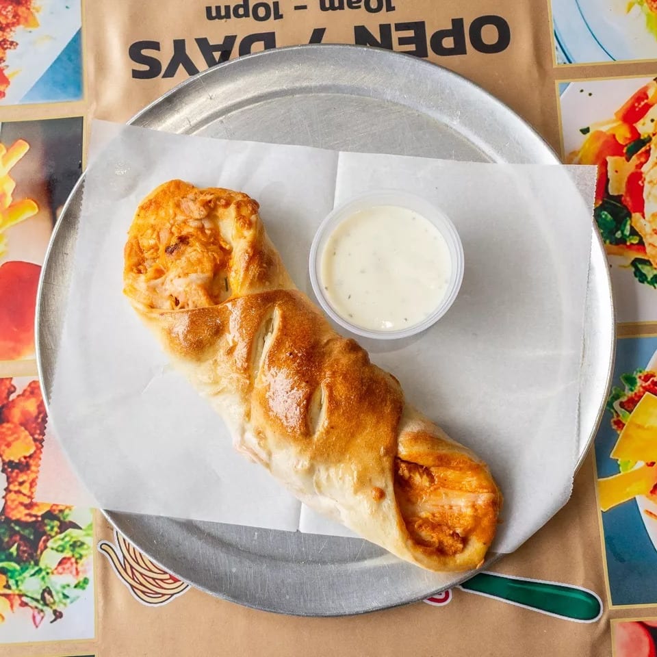 A golden-brown calzone with a side of white dipping sauce on a metal tray over a menu.