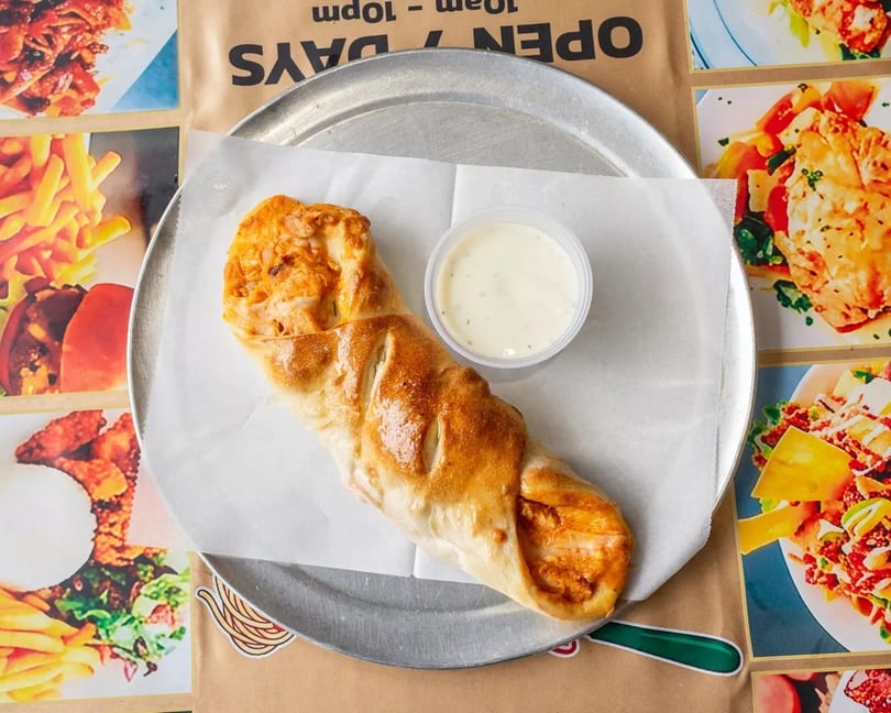 A golden-brown calzone with a side of white dipping sauce on a metal tray over a menu.