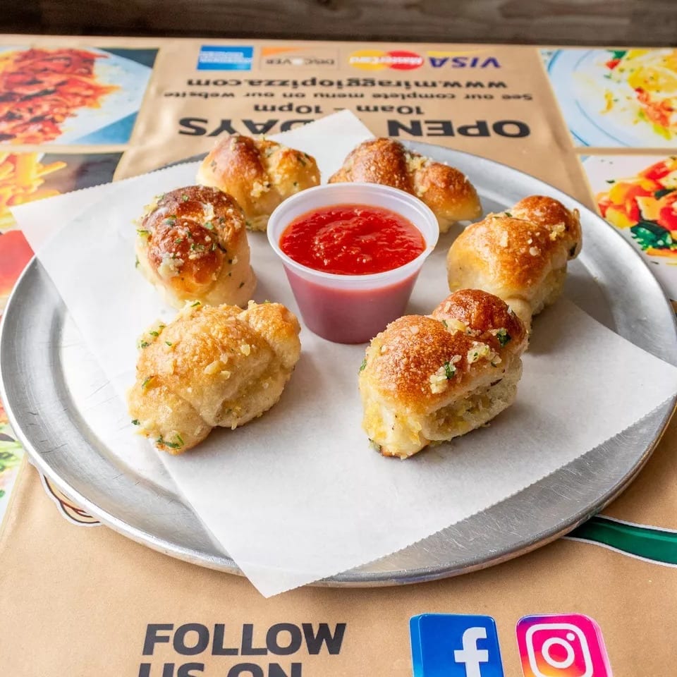 Six golden-brown garlic knots with herbs and a side of red marinara sauce on a metal tray.