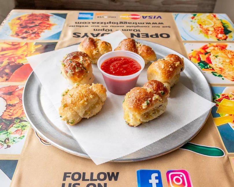 Six golden-brown garlic knots with herbs and a side of red marinara sauce on a metal tray.